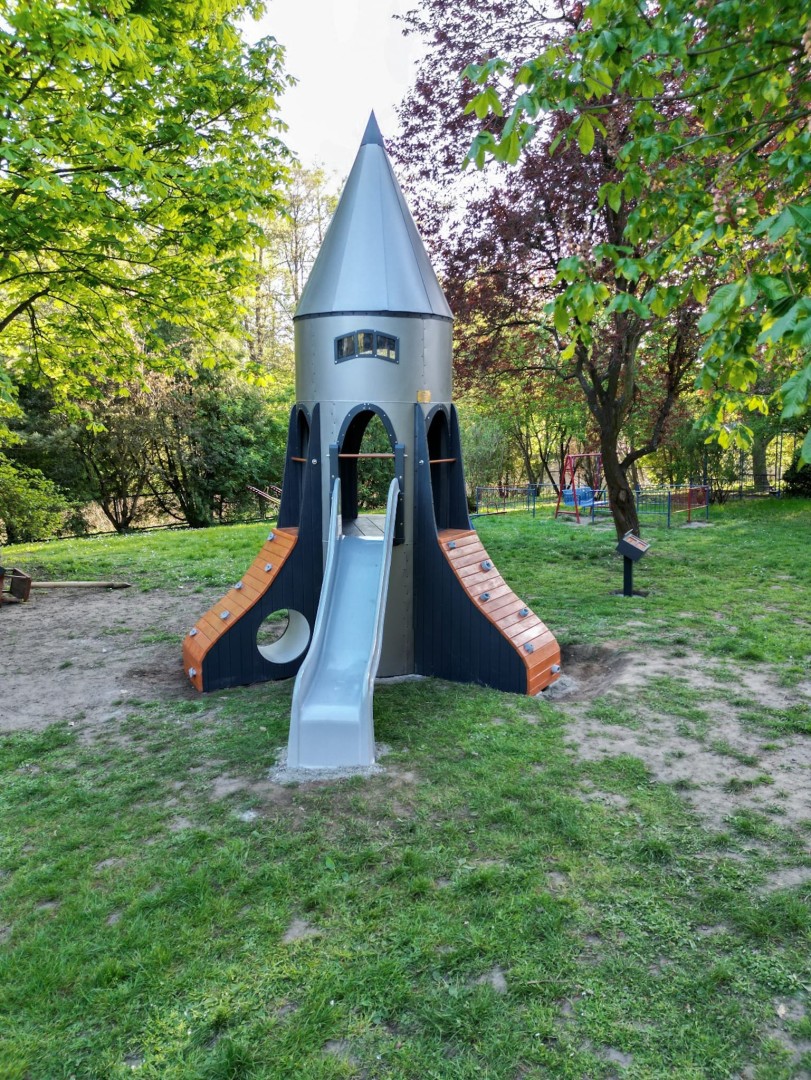 Plac zabaw z konstrukcją w kształcie rakiety, wyposażoną w zjeżdżalnię i ścianki wspinaczkowe, ustawiony na trawiastym terenie wśród drzew.